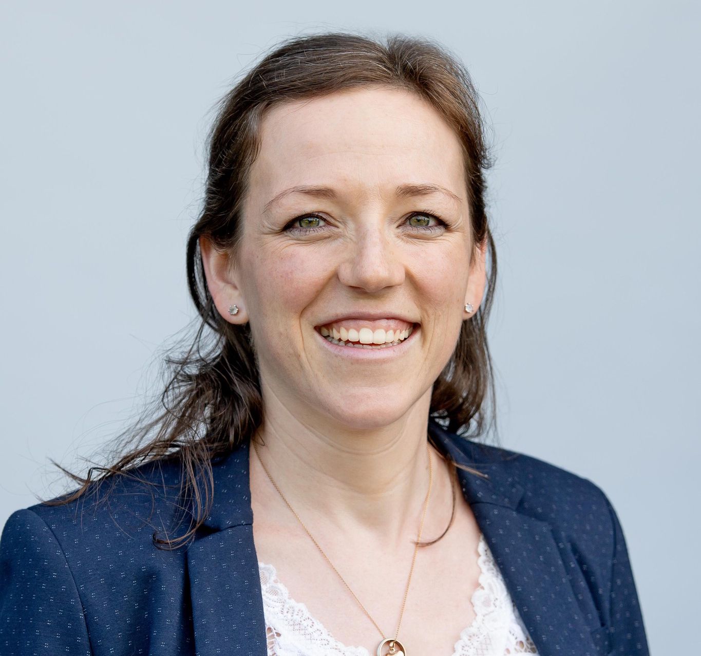 Femme souriante portant une veste bleue et une robe blanche en dentelle sur fond bleu clair.