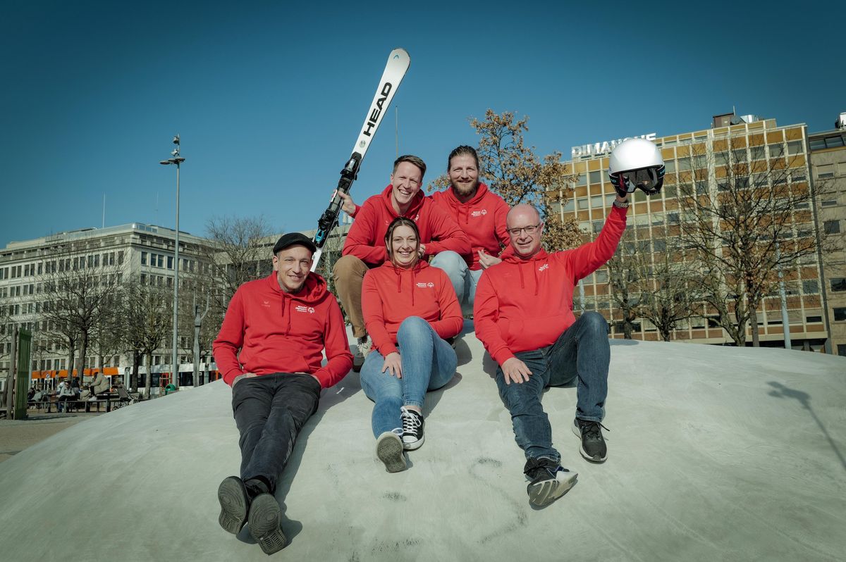 Cinq personnes en vêtements rouges sourient, assises sur une colline à Plainpalais, Genève. L’une tient un ski, une autre un casque.