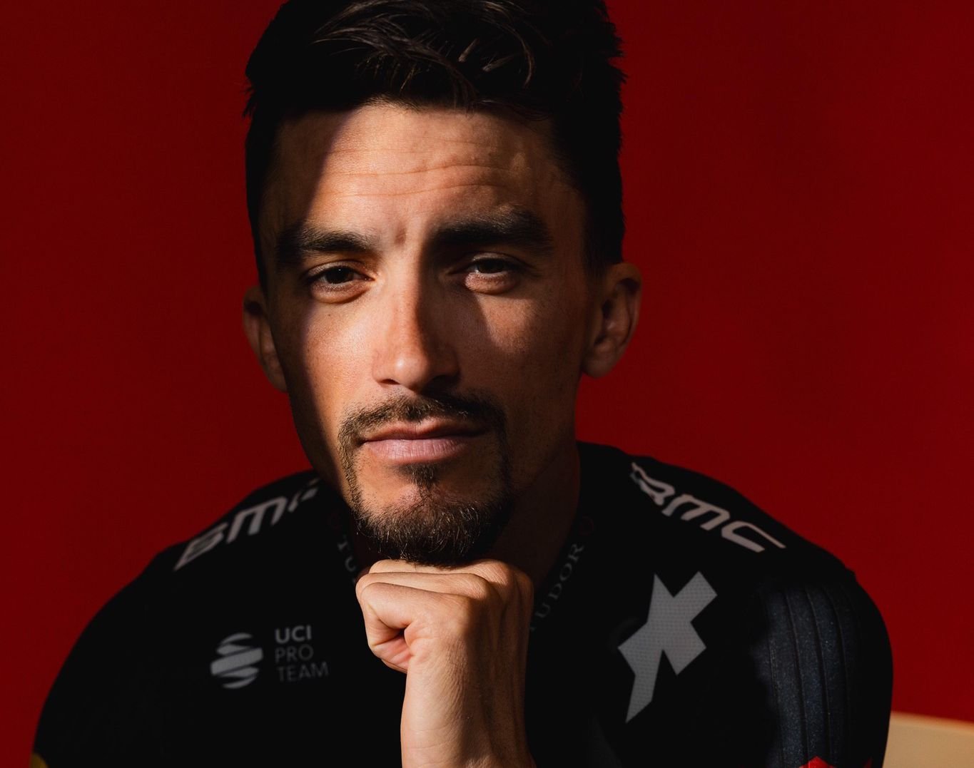 Julian Alaphilippe avec son nouveau maillot Tudor, assis devant un fond rouge.