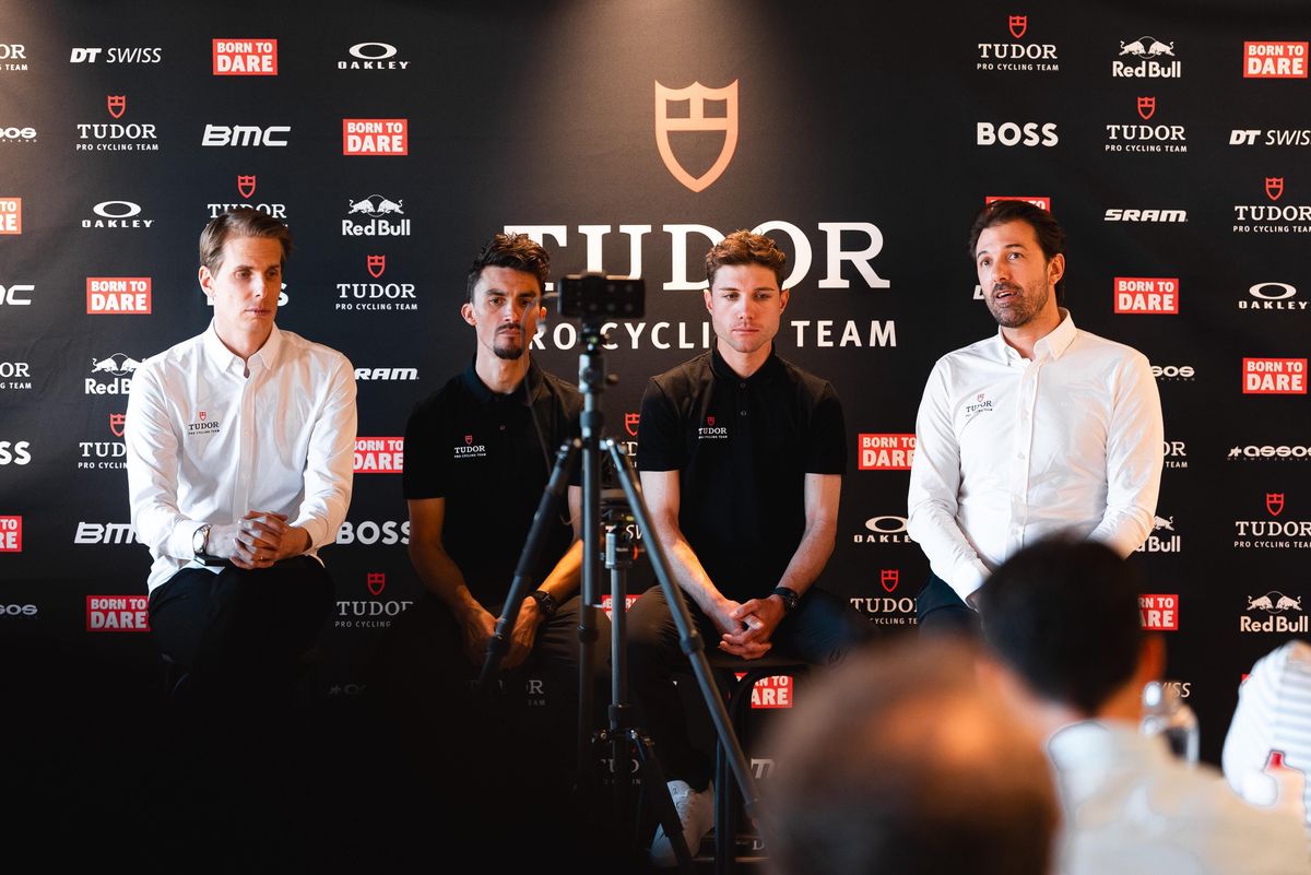 Quatre hommes assis sur une scène devant une bannière du Tudor Pro Cycling Team, lors d’une conférence de presse.