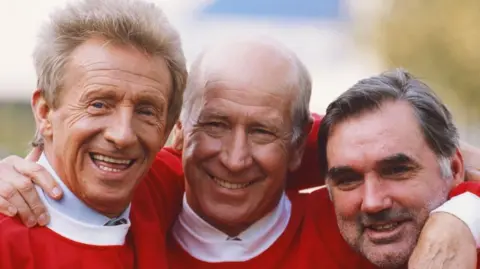 Denis Law avec Bobby Charlton et George Best, souriant à la caméra.