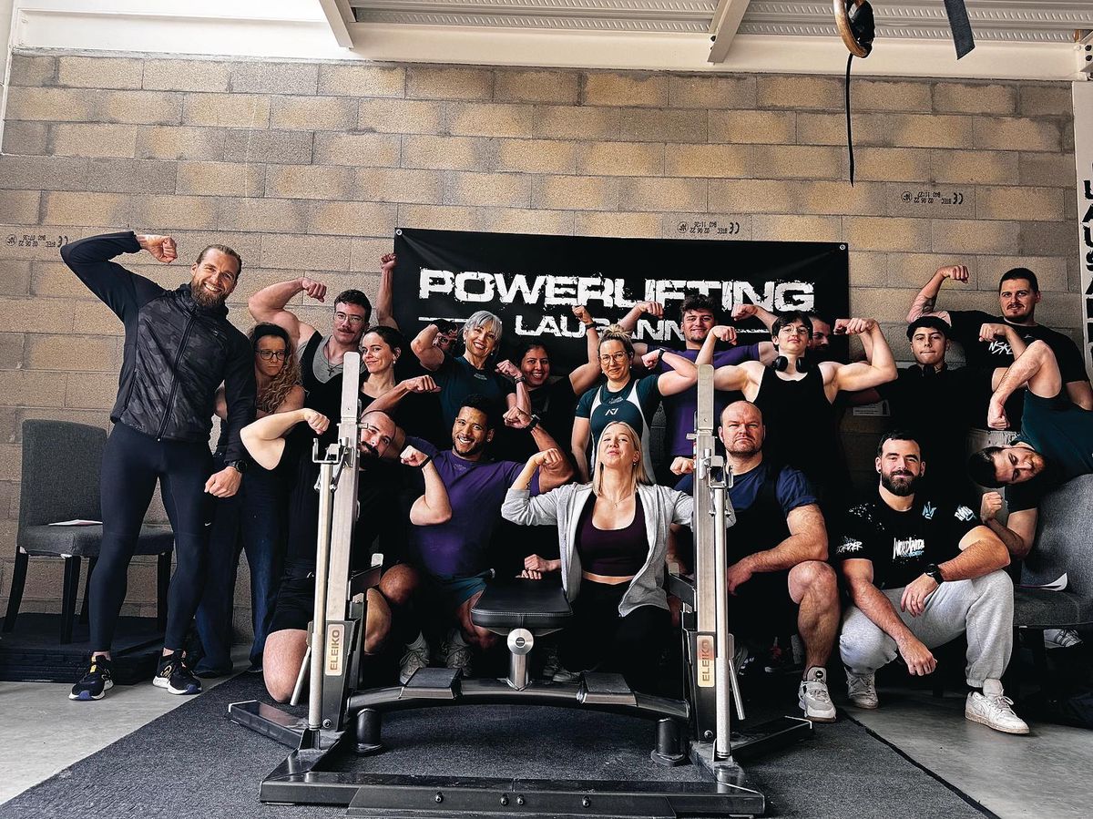 Groupe de personnes posant fièrement dans une salle de gym avec une bannière ’Powerlifting Lausanne’ à l’arrière-plan, montrant leurs muscles.
