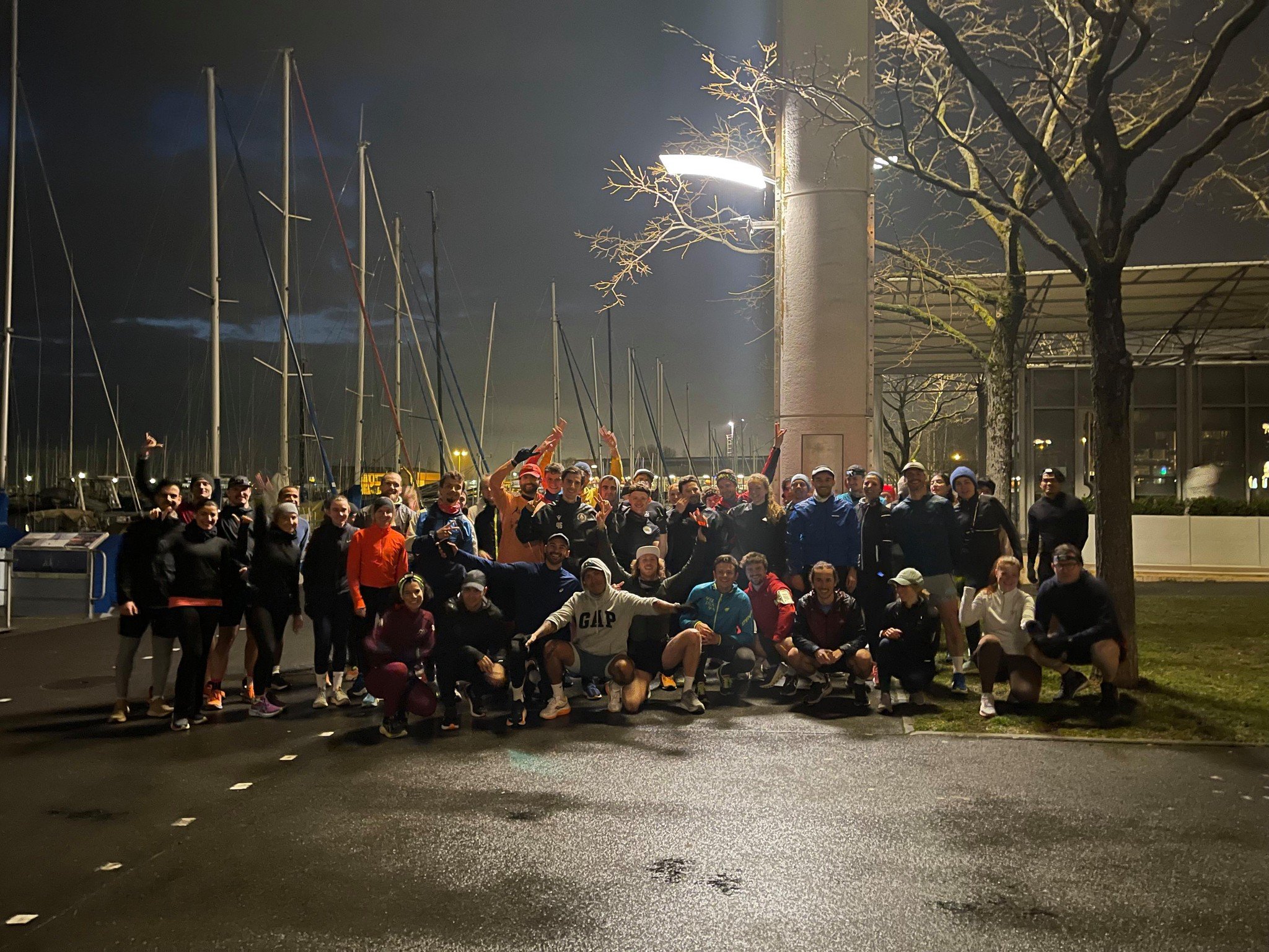 Groupe de personnes posant devant un port de plaisance la nuit, sous des lampadaires avec des voiliers à l’arrière-plan.